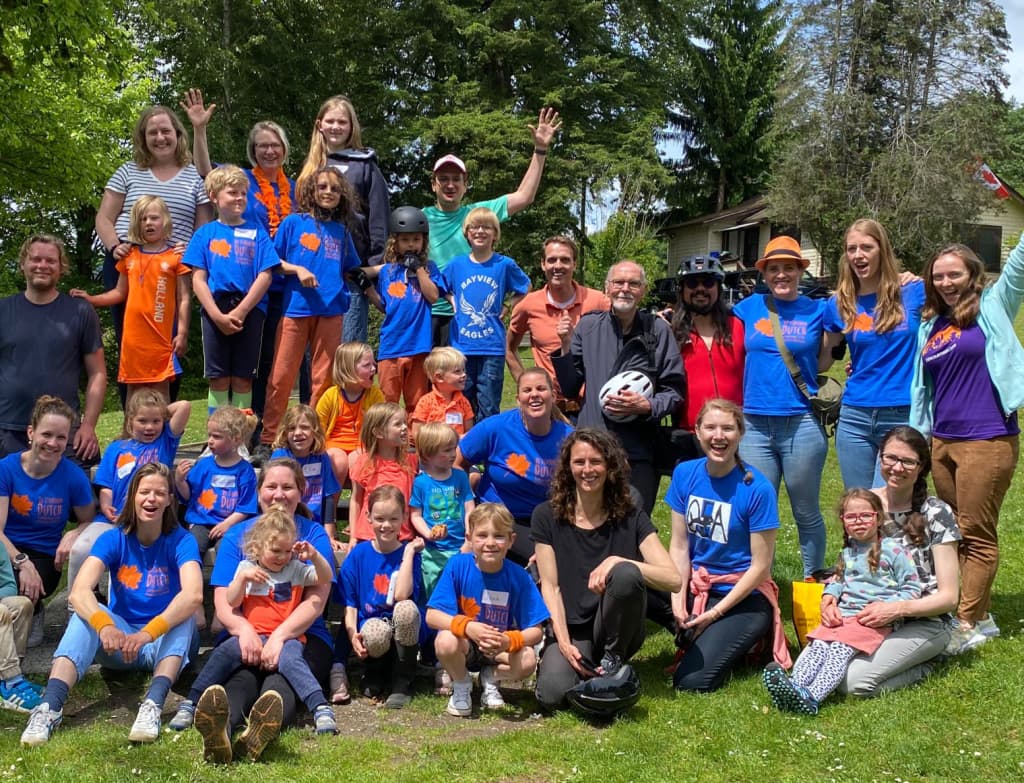 A large group of people, including children and adults, smiling and posing outdoors in a park. Many are wearing bright blue and orange shirts, suggesting a fun group event. Trees and grass provide a natural background